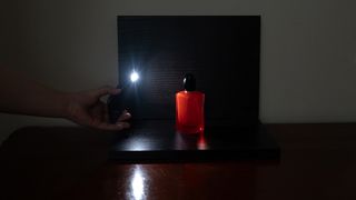 A person holding a phone torch next to the scene where a red perfume is placed in front and on top of a black ground