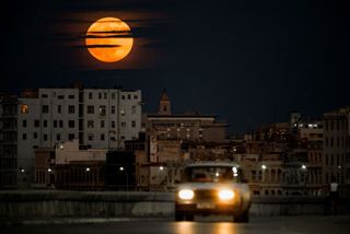 bright moon in sky partially shrouded by clouds shines above buildings with a car driving toward the camera in the foreground.