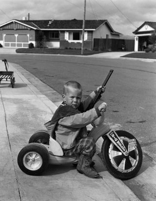 Black and white image of toddler on bike with gun. From the exhibition Suburbia. Building the American Dream