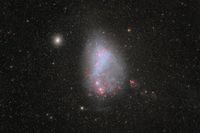 A black space background covered in stars has a central bright, egg-shaped galaxy with blue and red coloring. 