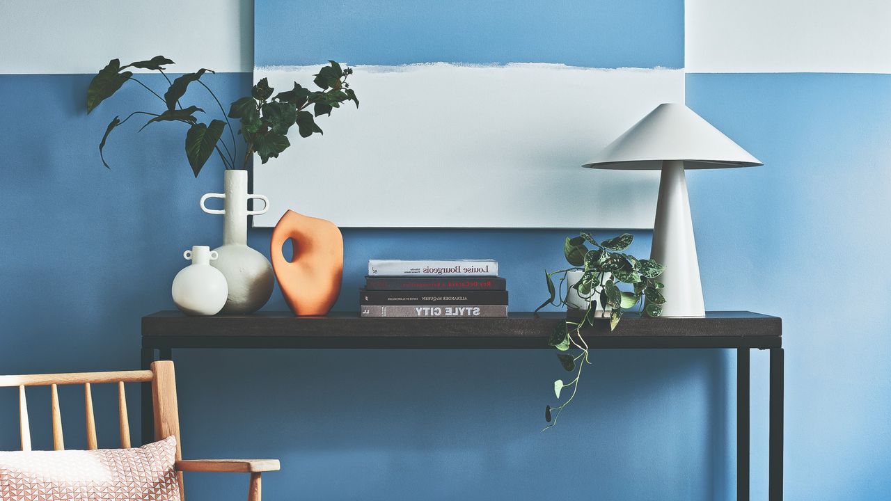 A console table against a two-tone white and blue wall with a sculptural white table lamp on top