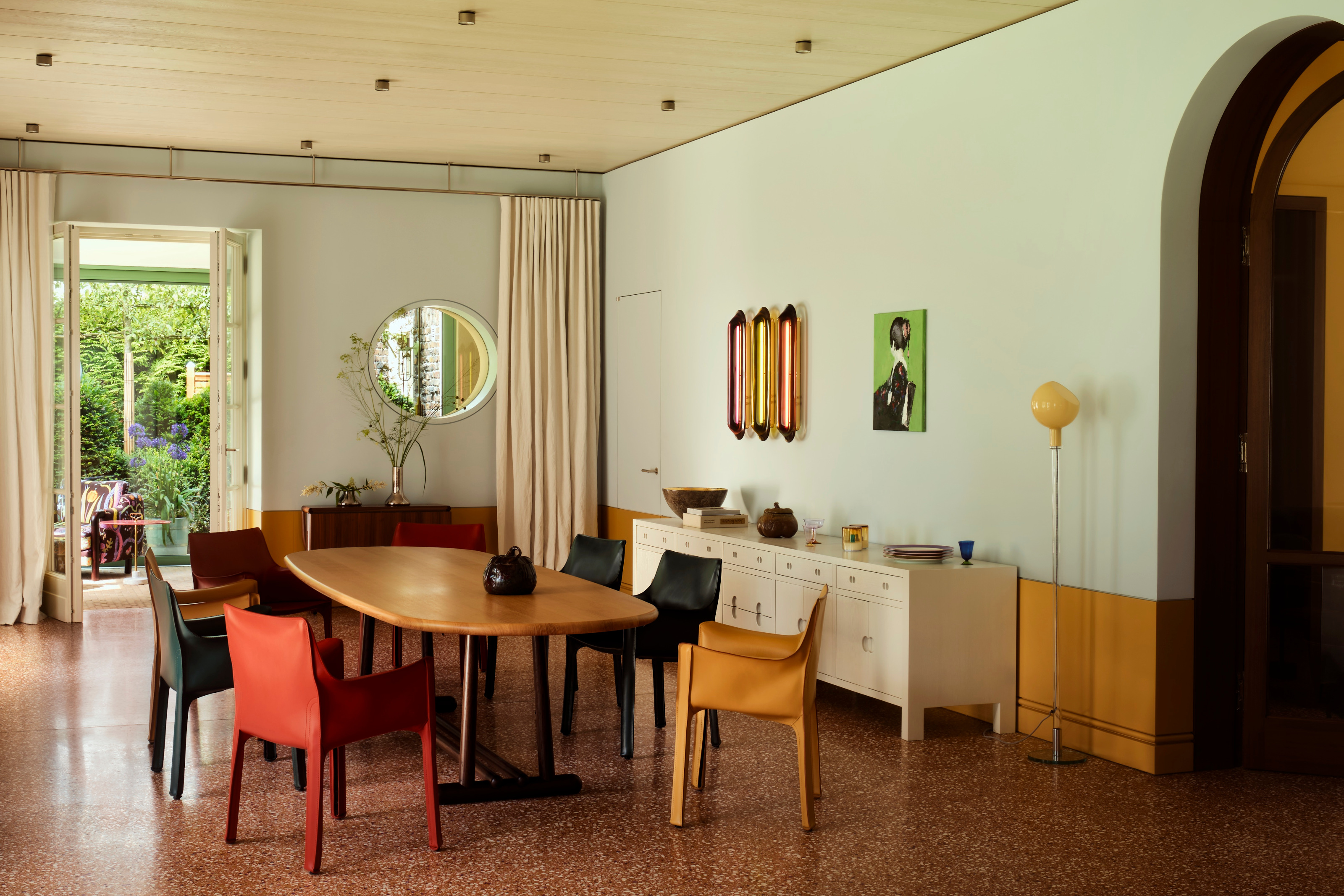 Dining area with oval wood tale, multicolor chairs, red terrazzo floor, pale blue walls, white sideboard and multicolor wall lights