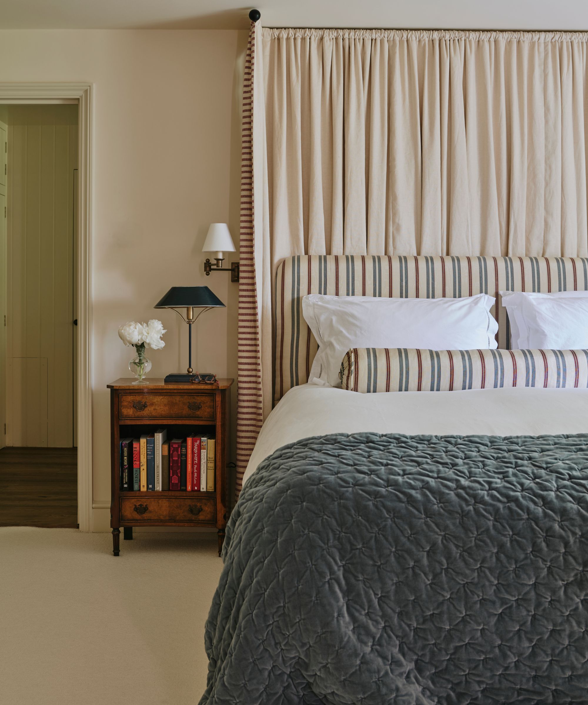 cozy neutral bedroom with a bed canopy, striped headboard and matching pillow, blue velvet quilt, vintage nightstand styled with books, a lamp and wall sconce