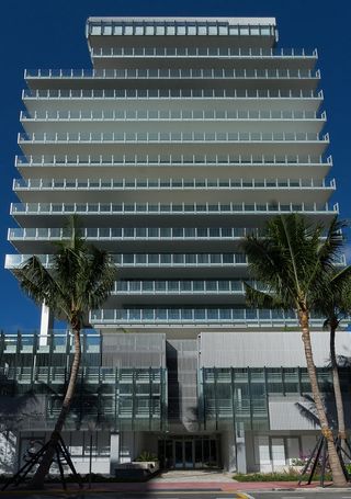 Front view of Rene Gonzalez’s crystalline tower. Building with glass banisters on the balcony's and palm trees in front of it.