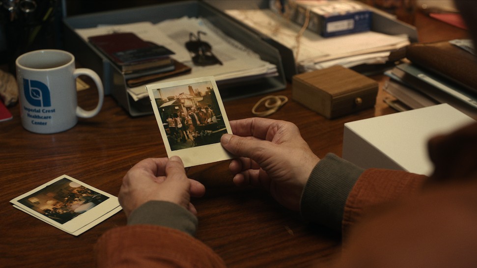 A person's hands hold up a square instant photo, showing a group of people outdoors, with other similar photos and office items on a wooden desk.