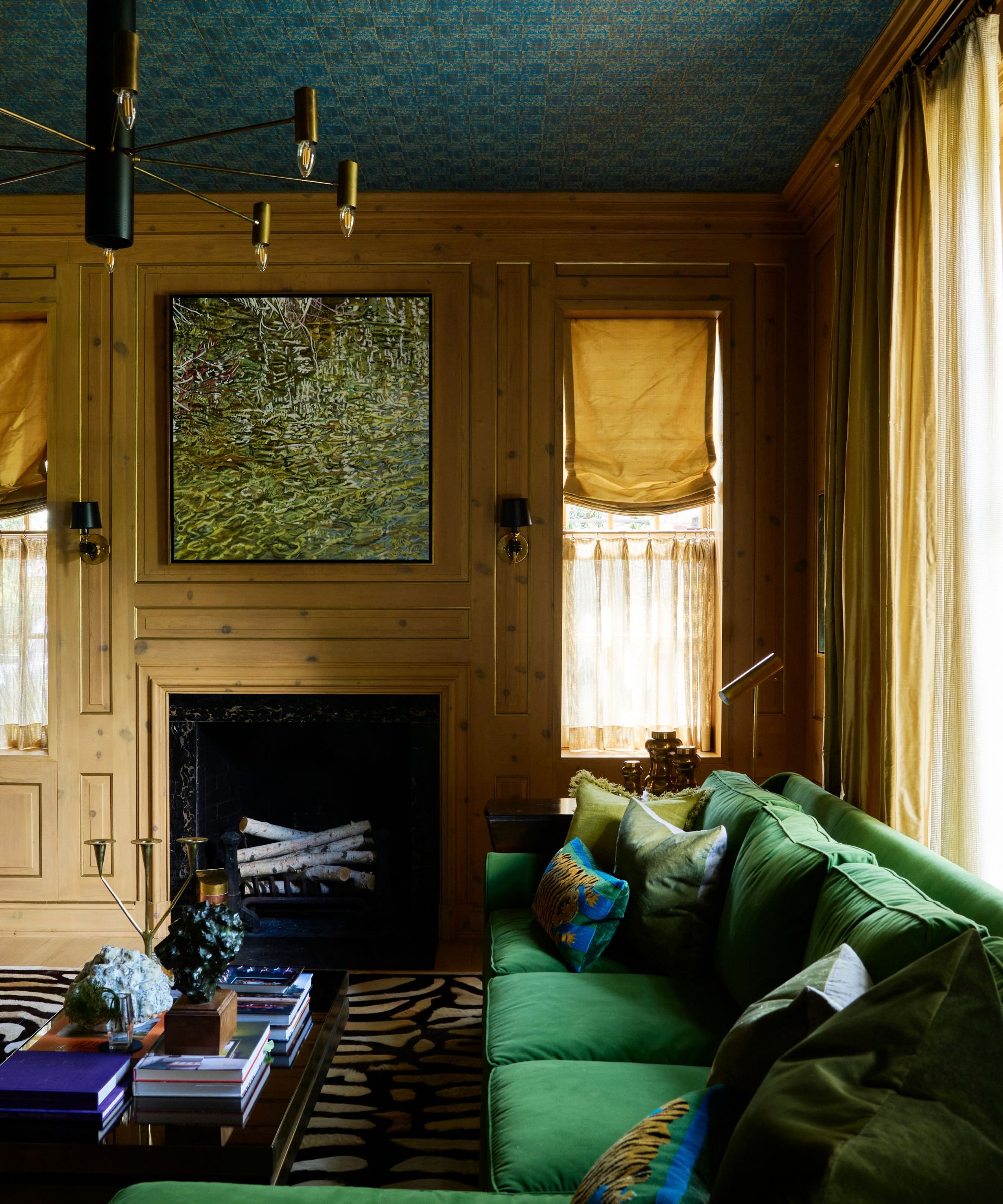 A large library room in a traditional home with natural wood wall paneling, a teal wallpapered ceiling, a dark green sofa with blue and green cushions, mustard roman blinds, a green artwork above the fireplace, and a black and cream patterned rug.