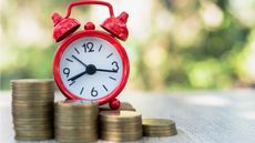 A red alarm clock sits atop stacks of coins of varying heights.