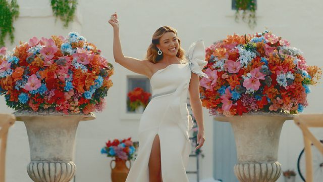 Dannii Minogue, wearing a white wedding dress, snaps her fingers and smiles as she hosts the 2024 I Kissed a Girl finale