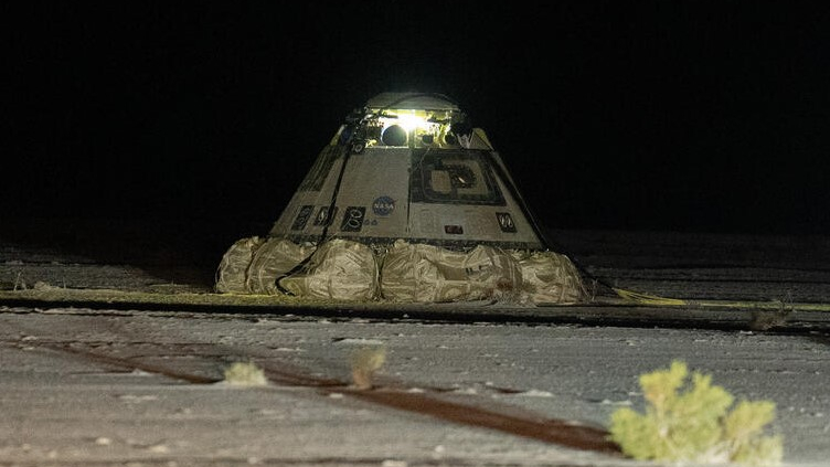 A burnt grey cone-shaped capsule lies on the desert sand surrounded by bushes.