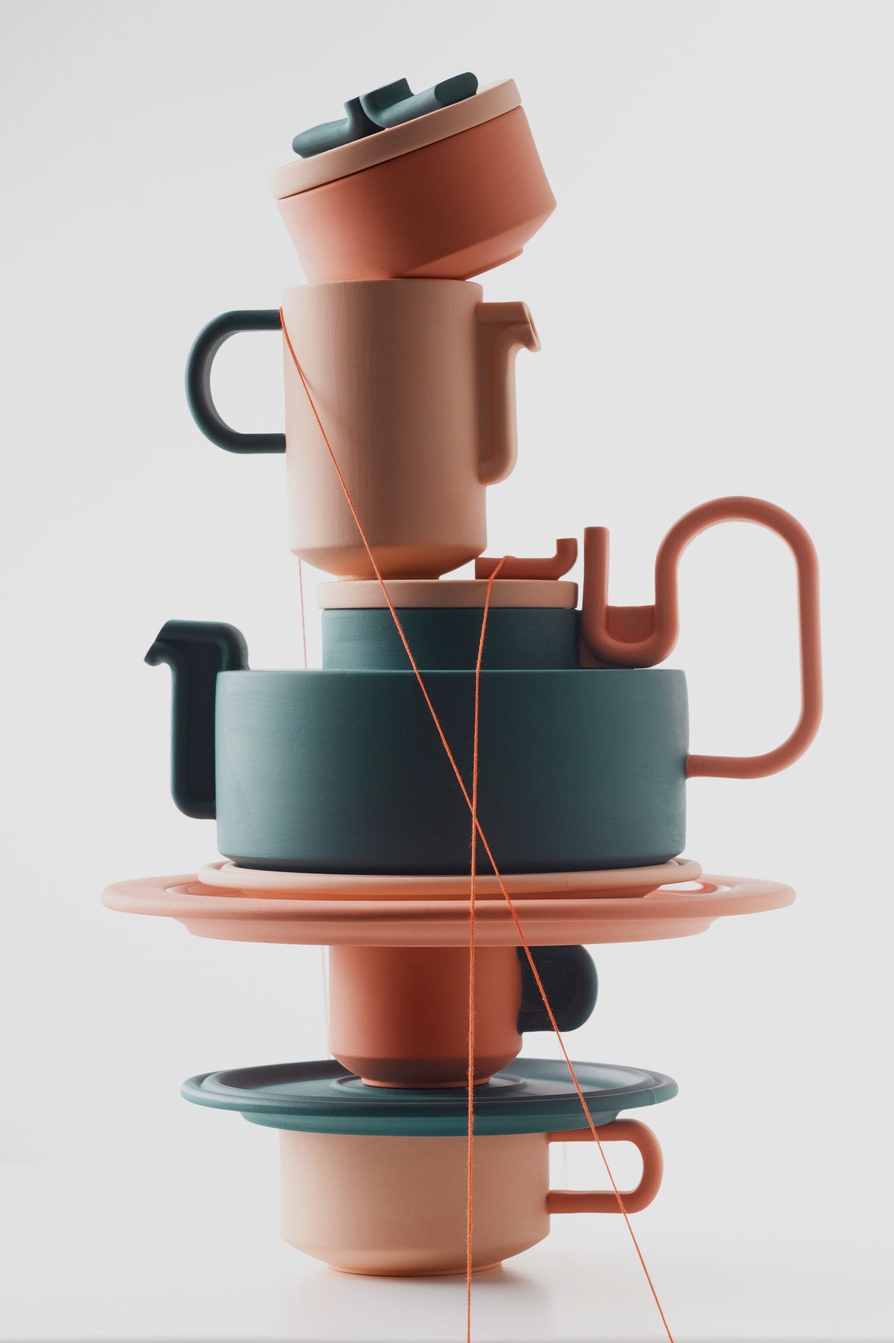 A series of cute, terracotta and muted petrol green cups, mugs, and saucers are assembled into an abstract composition kept by orange wire.