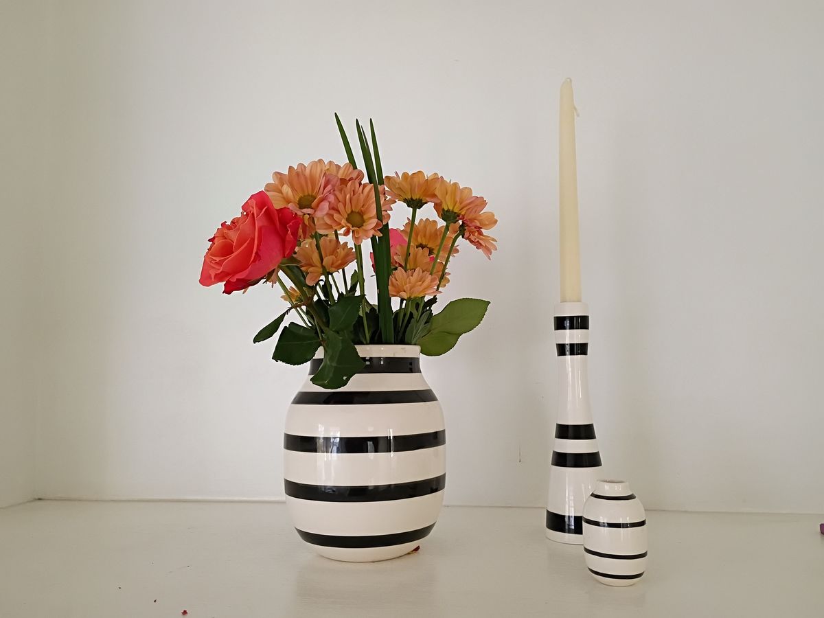 three white ornaments with black stripes on a white table in front of a white wall, with flowers in the vase and a white candle in the candle holder