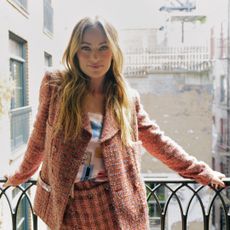 Olivia Wilde stands on a balcony wearing a tweed Chanel suit including wide leg pants and a blazer