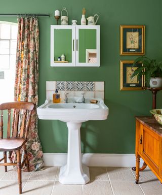 Traditional bathroom with green painted walls, white sink, cream stone flooring, wooden cabinet, dark wooden chair, floral curtains, white wall mounted cabinet with mirrored doors above sink, framed paintings on walls.
