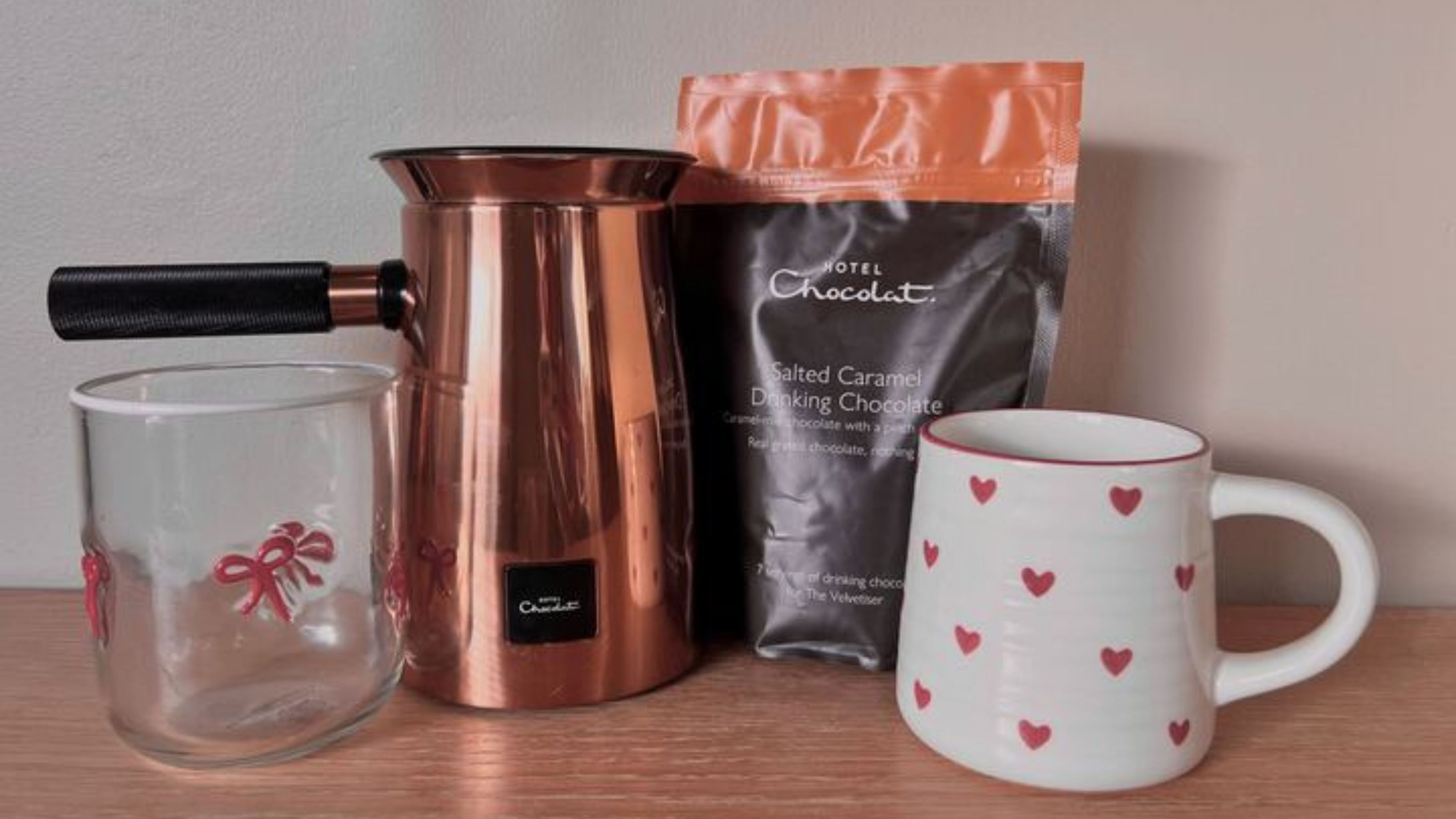 A Hotel Chocolat Velvetiser with a bag of salted caramel drinking chocolate, a white and red heart-patterned mug, and a glass with red bows. 