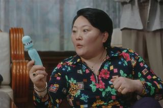 A middle-aged Korean woman with a surprised expression holds a light blue massager, in 'A Virtuous Business.'