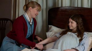 Pregnant Paula lies in bed while Rosalind sits beside her and holds her hand reassuringly.