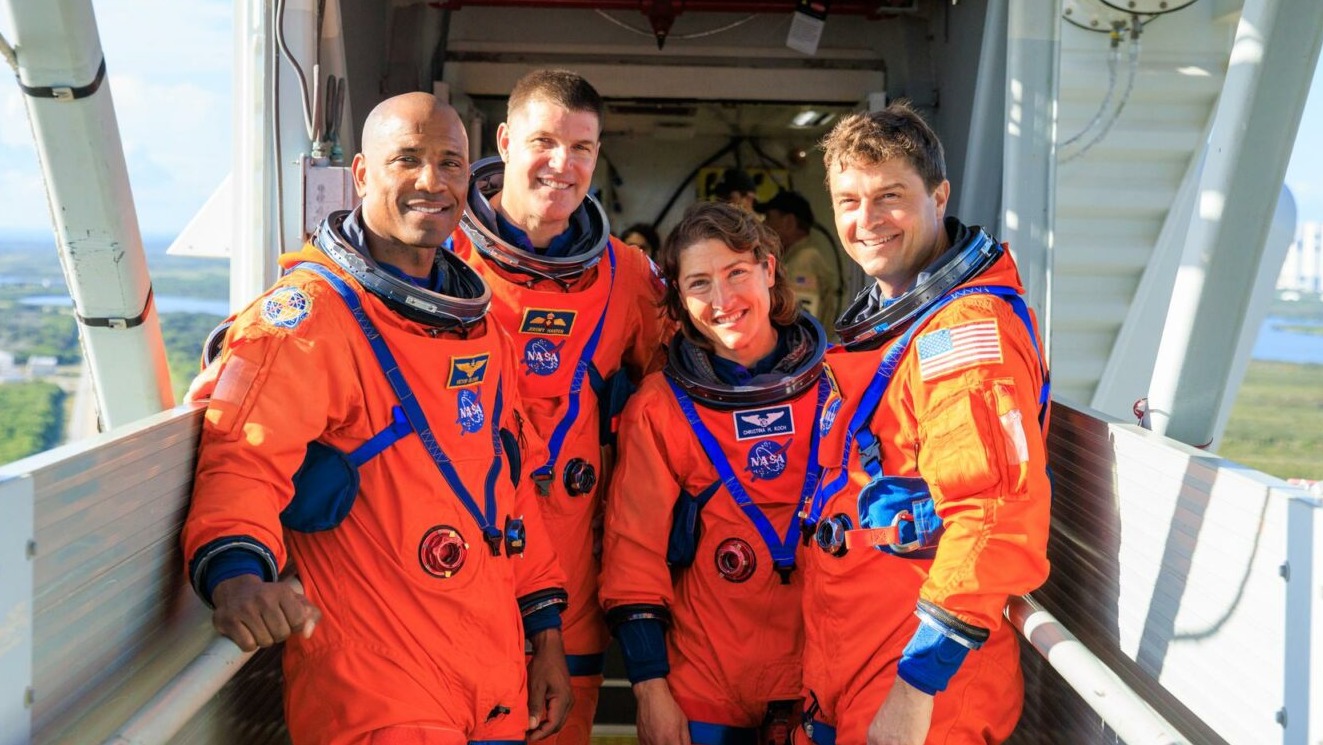 four people in spacesuits standing in a group and smiling at camera. behind is distant view of water and grass