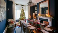 A blue dining room decorated for Christmas with a large laid table, large Christmas tree, presents, taper candles and a decorated white mantelpiece with candles, foliage, and a large gold mirror above. 