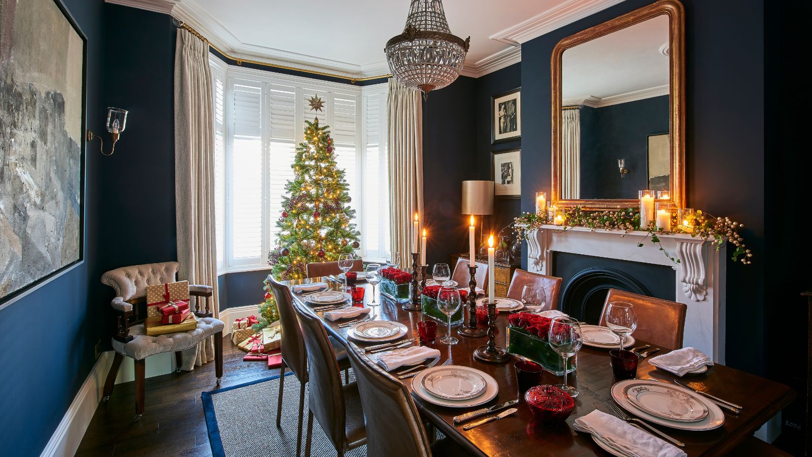 A blue dining room decorated for Christmas with a large laid table, large Christmas tree, presents, taper candles and a decorated white mantelpiece with candles, foliage, and a large gold mirror above. 