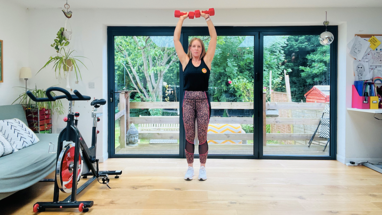 Personal trainer Maddy Biddulph performs the first part of an Arnold press in her well-lit living room. She wears a black vest, leopard-print leggings and white sneakers. She is standing and holding a pair of light dumbbells overhead, with her arms extended. Behind her is a window showing a leafy garden and next to her is an exercise bike.