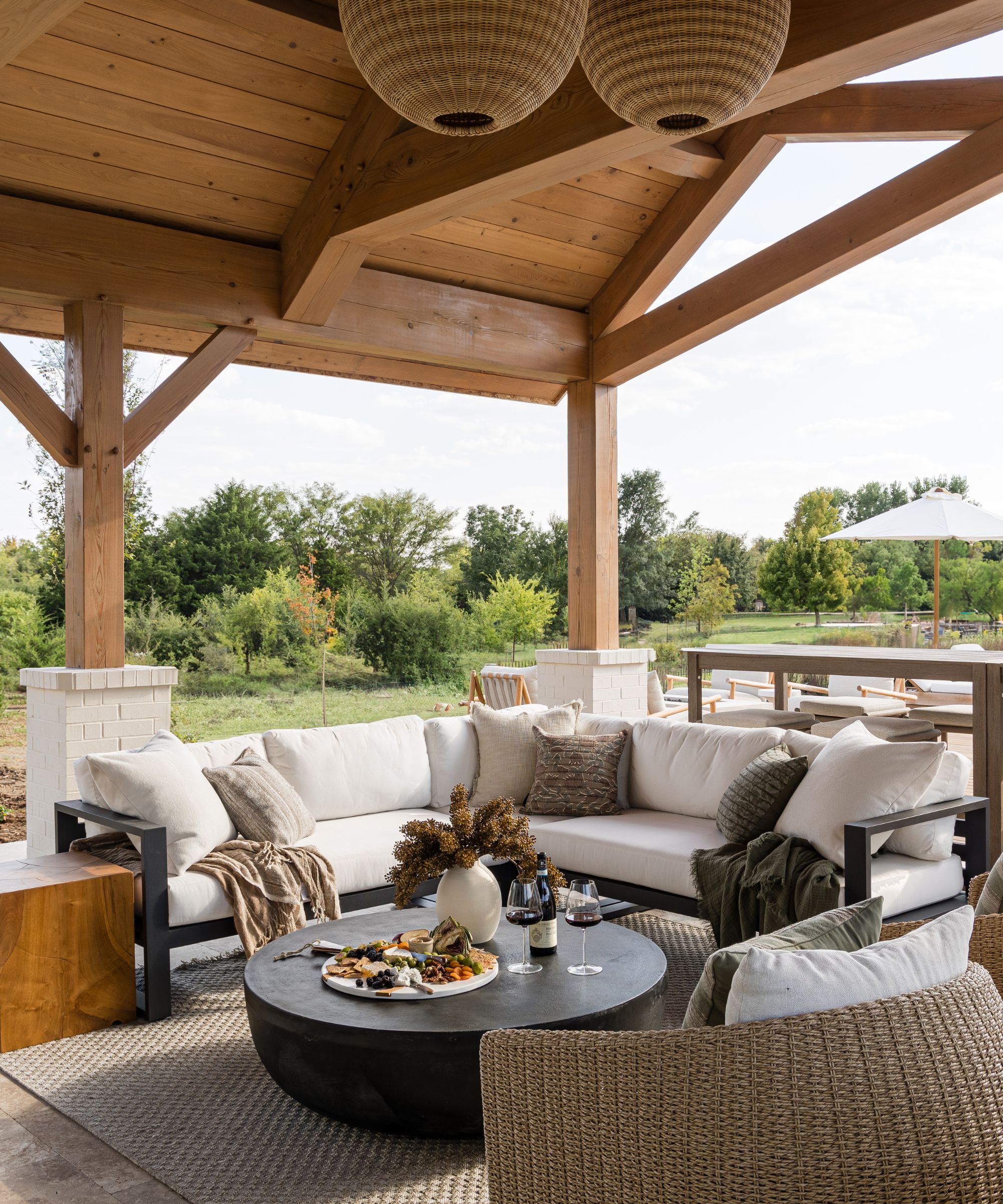 outdoor living room under a wooden roof with a corner sofa, black wooden coffee table, wicker chairs, cushions and throw blankets
