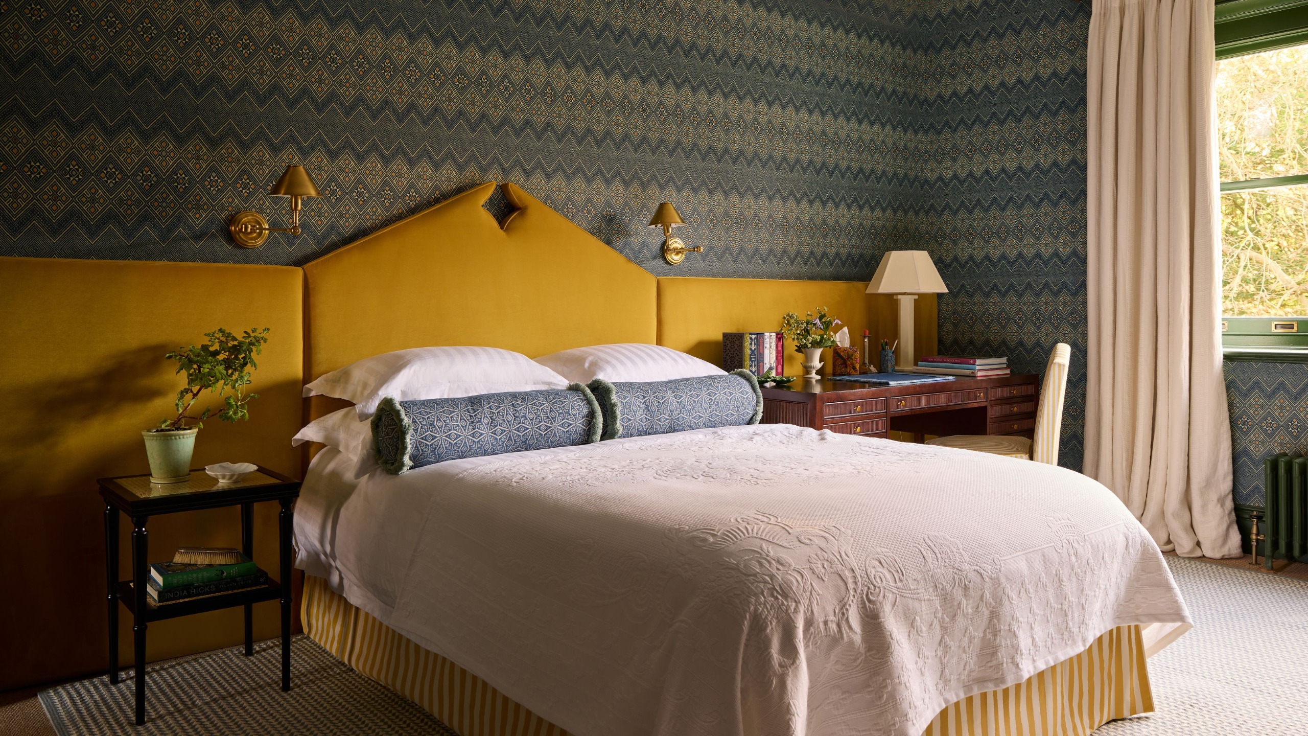 A bedroom with a dark blue patterned wallpaper, an ochre headboard elongated the full length of the wall, neutral bedding with dark blue bolster cushions, cream curtains, and a wooden desk.