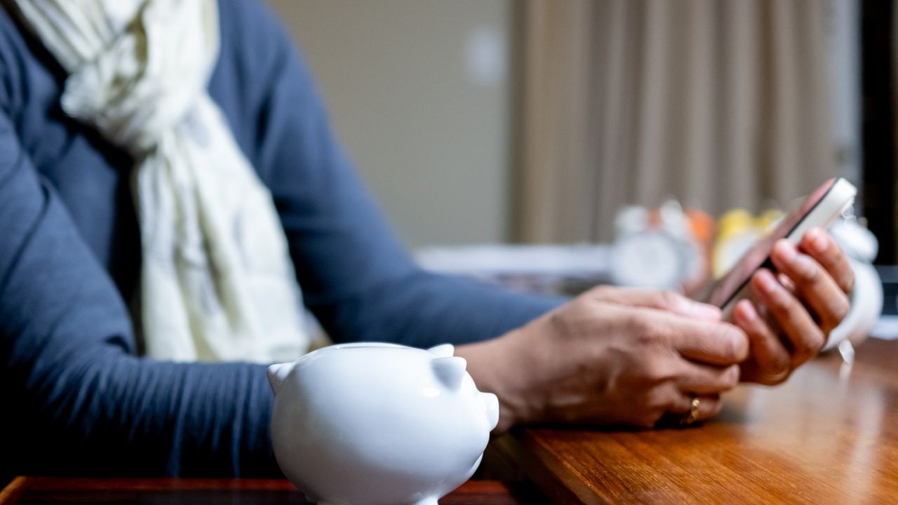 Woman looking at her smart phone at desk with a piggy bank sitting beside her