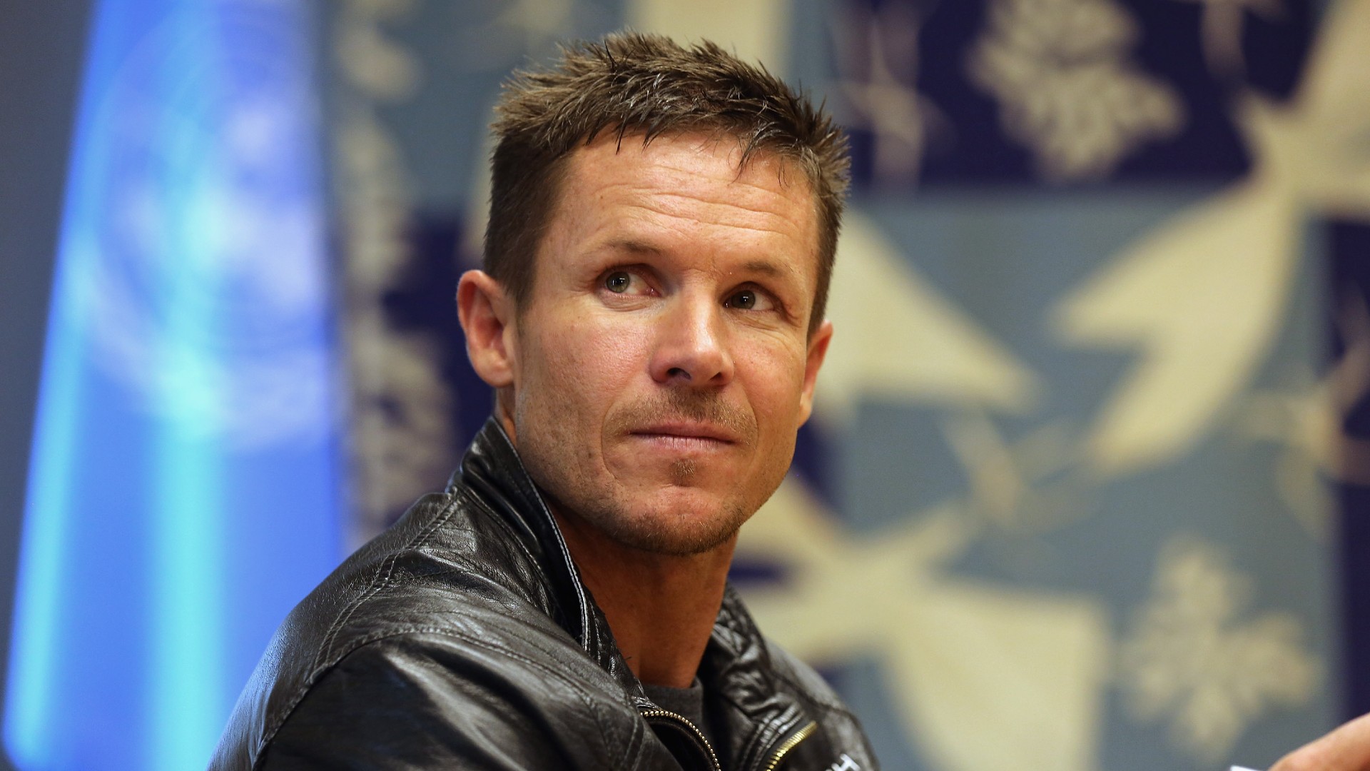 a man with short hair in a leather jacket smiles for a portrait in front of the united nations&#039; flag