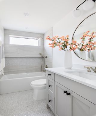 White and grey tiled bathroom styled with pink blooms in a white vase, in front of a round mirror