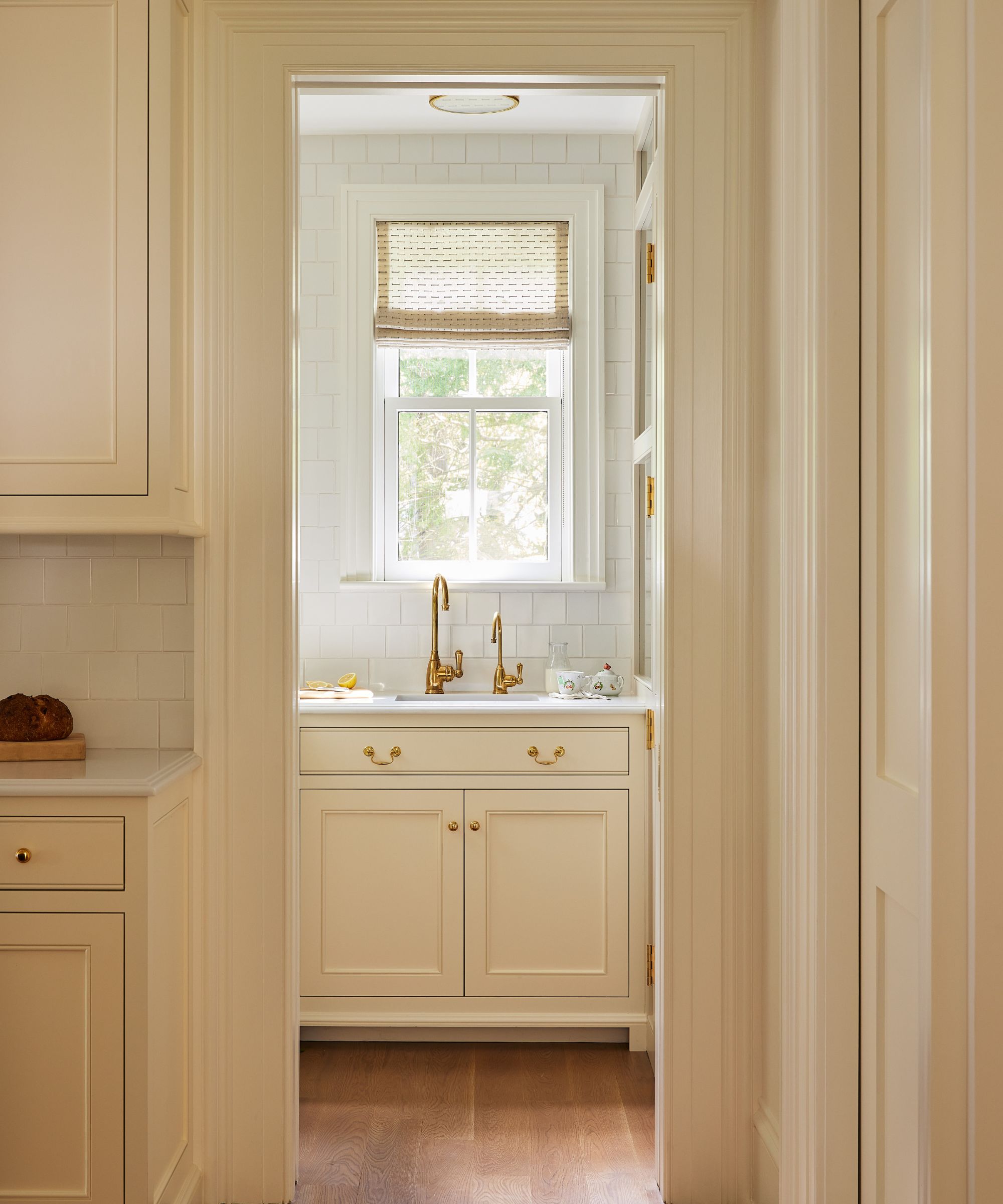 Kitchen looking into scullery with white tiles, cream cabinets and gold hardware and tap