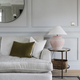 a neutral look living room with a cream sofa and velvet cushions, glass and metal side table, wall panelling and an oversized round foxed mirror