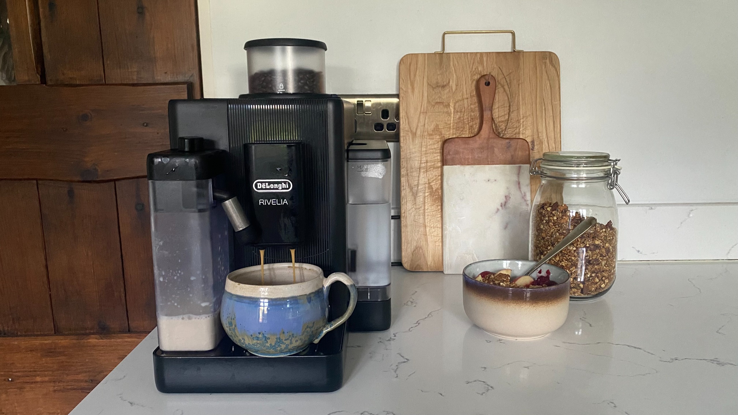 De'longhi Rivelia Start coffee machine making a flat white on a white stone kitchen counter with chopping boards and granola