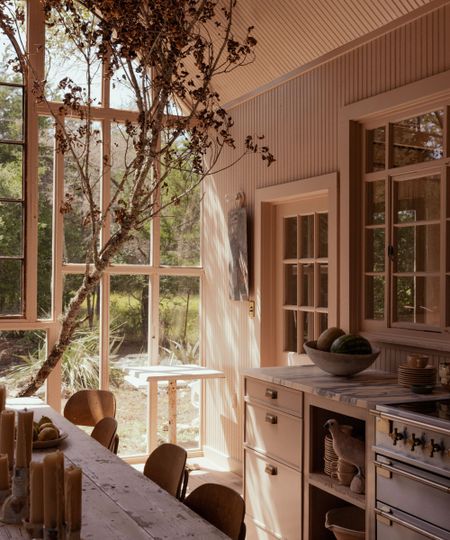 Light-filled kitchen with large glass doors, pink beige painted walls, wooden dining table with vintage wooden chairs, marble white countertops, and a silver range oven,