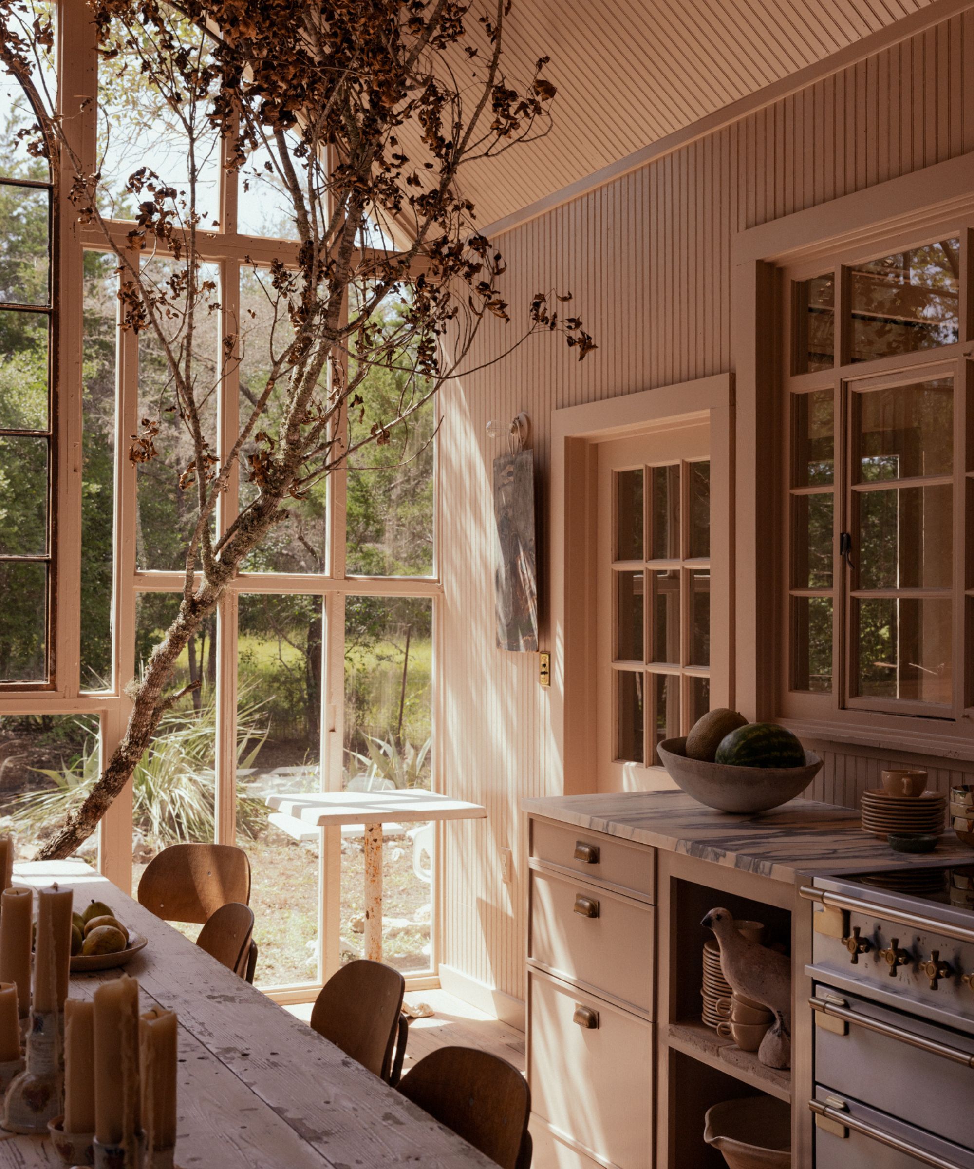 Light-filled kitchen with large glass doors, pink beige painted walls, wooden dining table with vintage wooden chairs, marble white countertops, and a silver range oven,