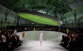 A fashion show featuring a ripple floor design in the center, with models walking around being watch by an audience