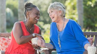 Two senior multi-ethnic woman, one Caucasian and the other African American, sitting on a porch enjoying each other's company. They are facing each other, laughing, talking and drinking coffee.