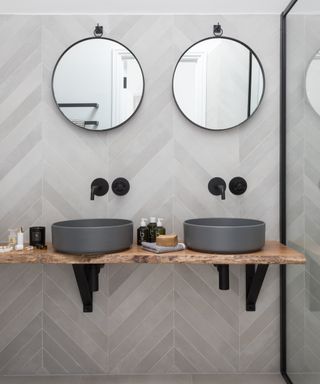 A bathroom idea with gray tiles behind a pair of gray sinks and mirrors.