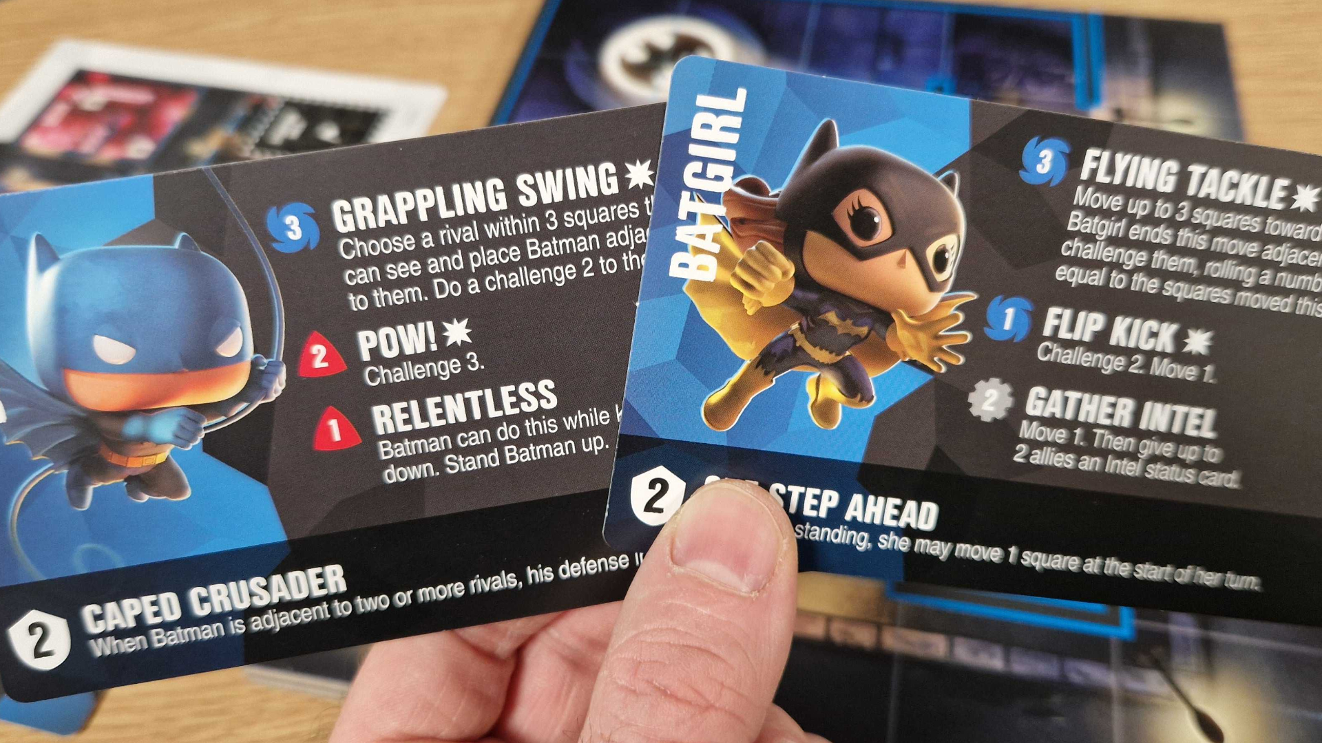 A hand holding Batman and Batgirl Funkoverse Strategy Game cards, with the board visible in the background