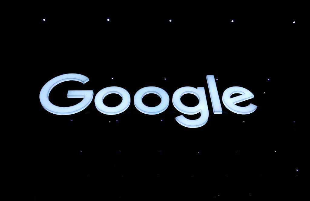 Google: A low-down shot of an illuminated pale blue Google logo, hanging from the ceiling in a pitch black room