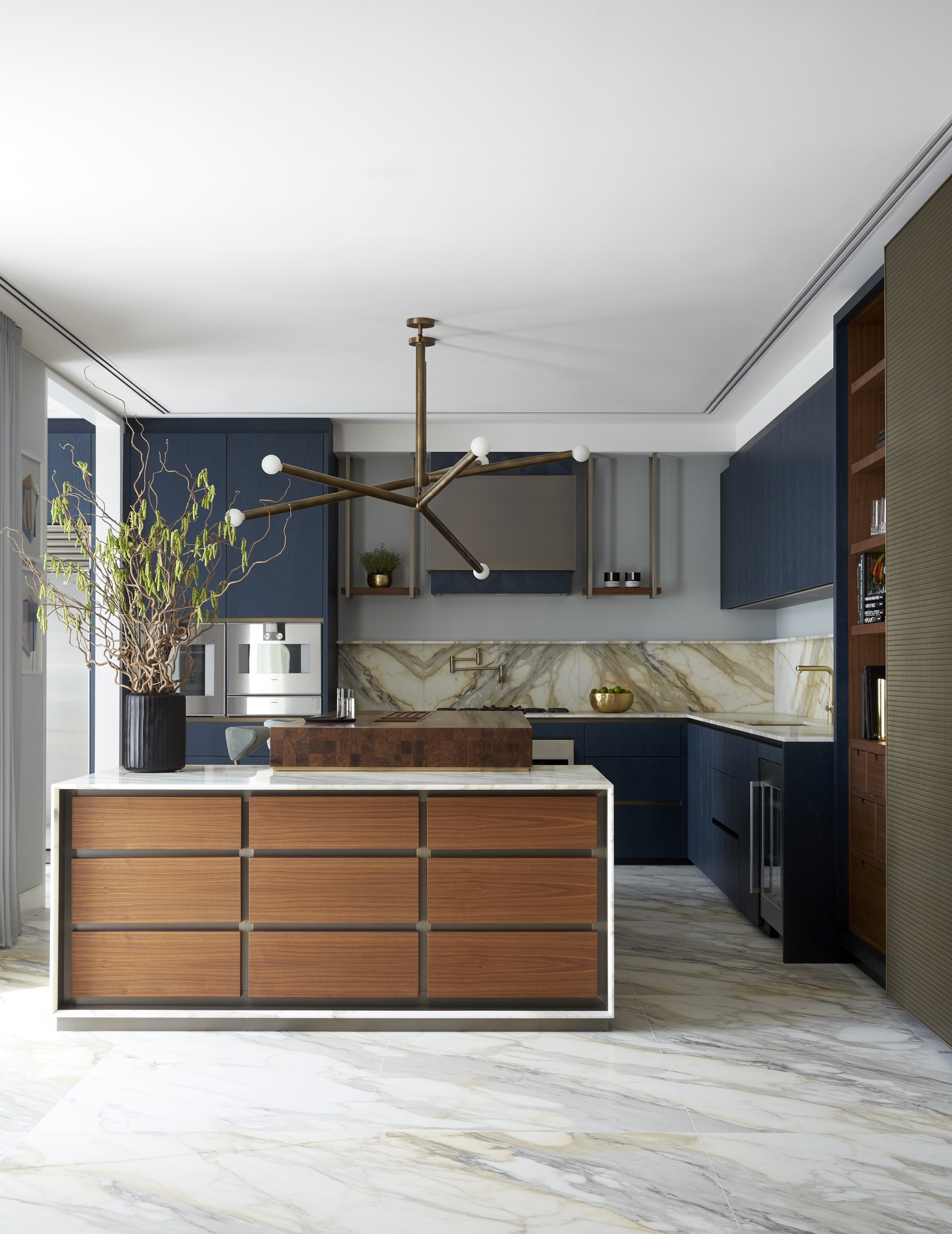 modern kitchen with marble flooring, timber kitchen island, dark blue kitchen cabinetry, marble backsplash, built-in appliances, and a vase with large foliage on the counter