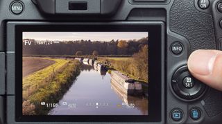 Rear LCD of Canon camera showing Fv mode activated while framing a canal scene. A finger is using the Quick Control Dial