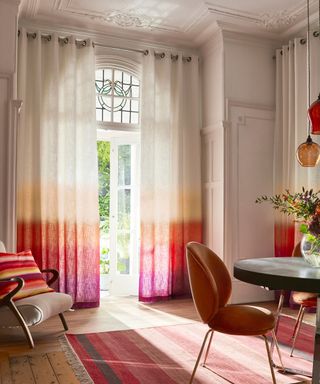 Large traditional dining room with colorful, modern design. Ombre pink, orange and white curtains, light wooden flooring, pink striped rug, rounded metal dining table with velvet beetle dining chairs