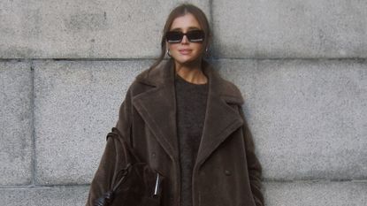 Woman standing against gray wall wearing brown sweater and brown teddy fall coat, black glasses, and gold hoop earrings.