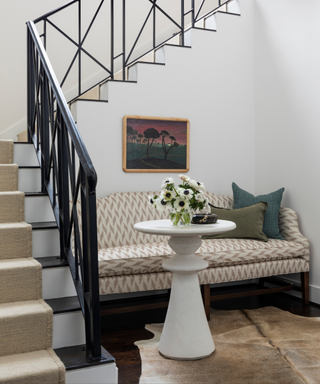 An entryway with a staircase and patterned bench, with a plaster-covered side table.