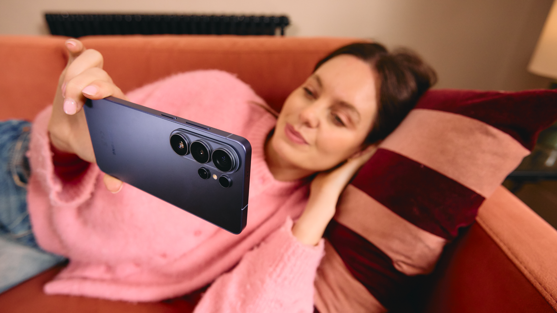 A woman smiles as she rests on the sofa, holding a Samsung Galaxy S26 Ultra in her hand