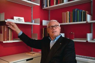 Dieter Rams in a suit with his righ hand reaching for a book on the white shelf which is against a pink wall