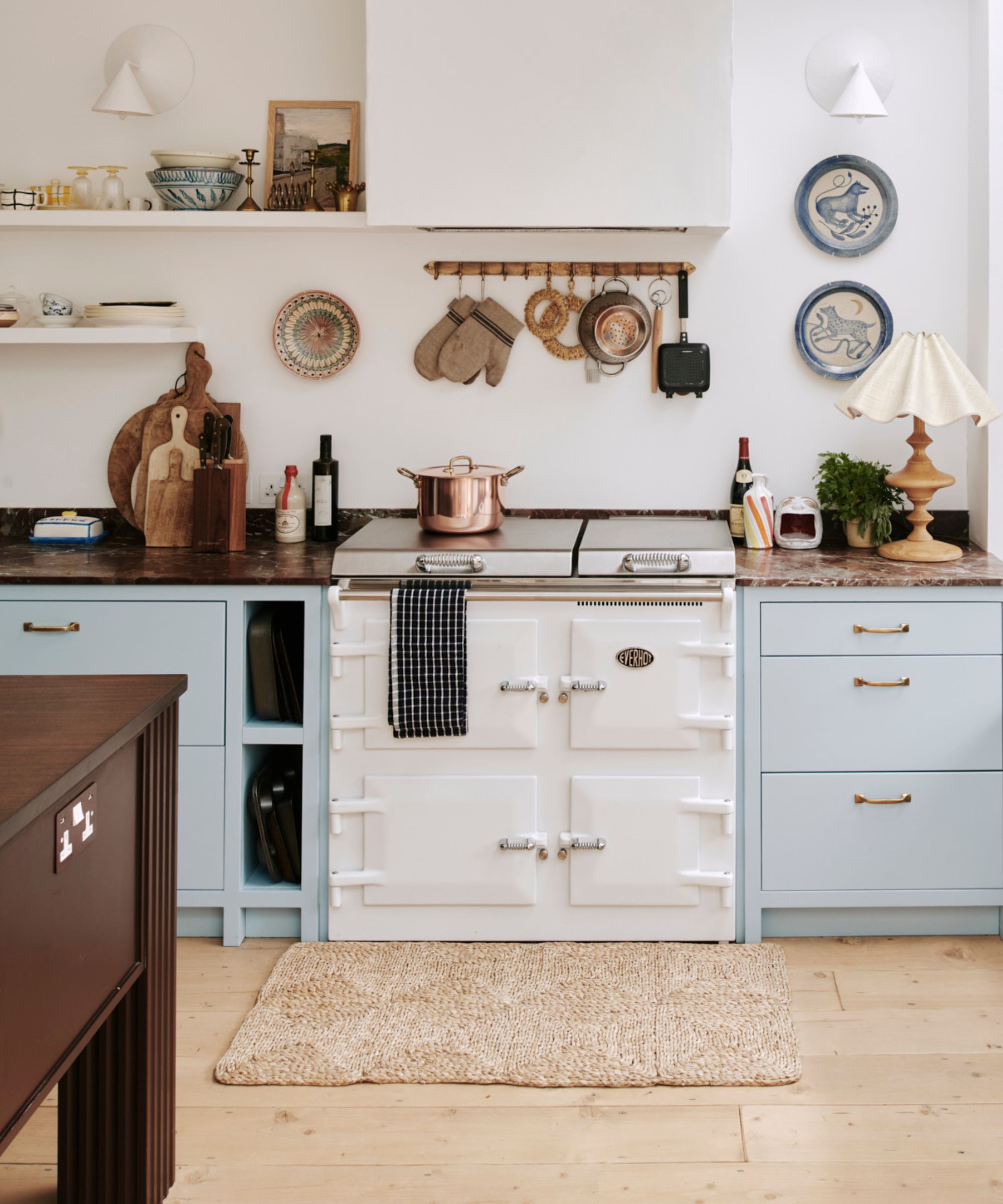 A cosy kitchen centres on a cream range cooker set between pale blue cabinets with brass handles. A copper pot sits on top. Open shelving, hanging utensils, and a cushioned window seat create a warm, lived-in atmosphere with soft, rustic charm.