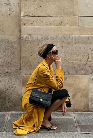 A woman wearing a black headband, gold earrings, a yellow trench coat layered over black capri pants, a black tank top, and black mules.