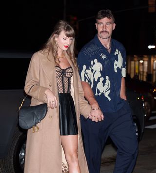 Travis Kelce wearing a blue printed shirt and blue pants holding hands with Taylor Swift wearing a tan trench coat and short black skirt with a sheer corset