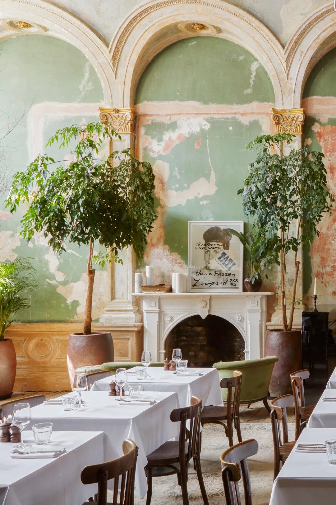 A romantic restaurant with raw, unfinished walls in green and pink tones boasts French bistro-style white table clothes and wooden chairs, thriving plants, and creative artwork on its arched walls.